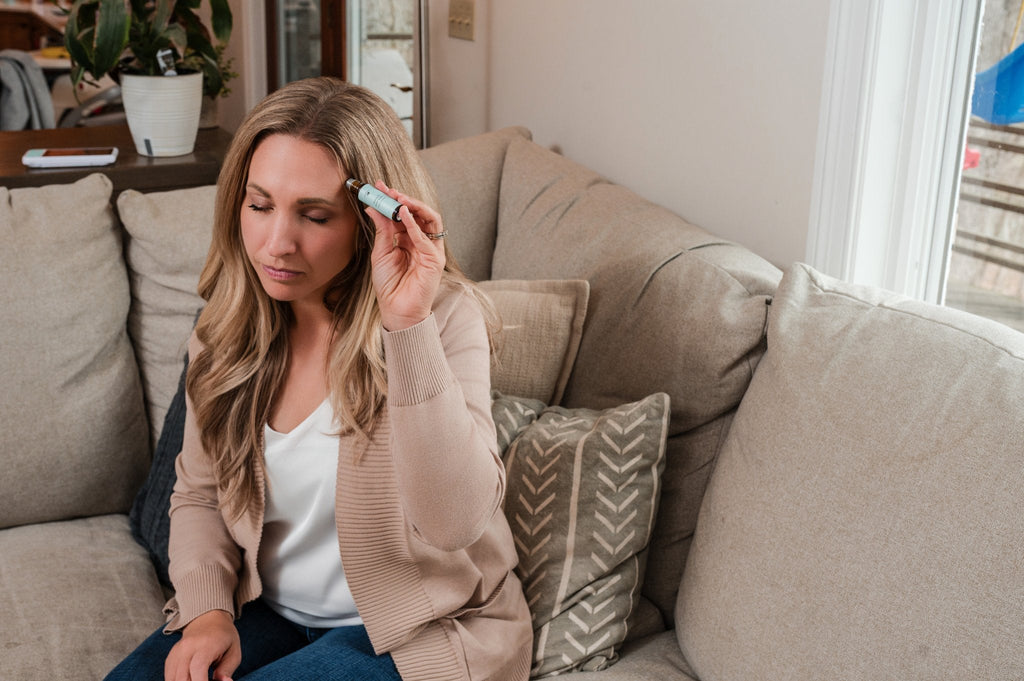 Woman sitting on a couch using a roll-on essential oil blend on her temple in a relaxed home setting, demonstrating natural aromatherapy for stress or headache relief.